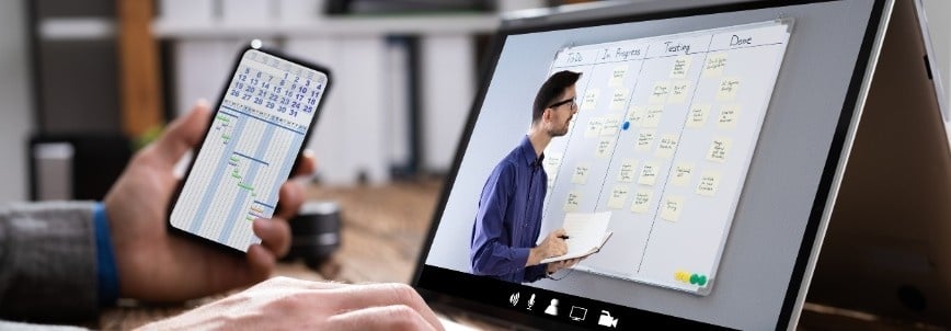 A person holds a smartphone displaying a project schedule, while watching a man present a task board with sticky notes on a computer screen during a virtual meeting for Rosemet CAPM training.