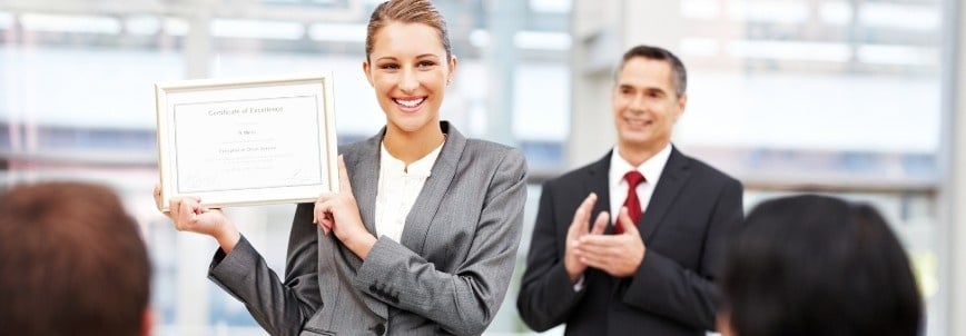 A woman in a gray suit smiles while holding up a certificate of excellence, possibly for meeting PMP certification requirements. She stands before an audience, with a man in a suit clapping in the background at this professional event.