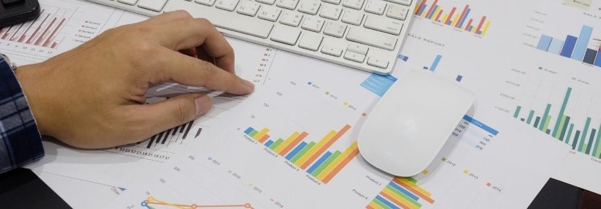 A person’s hand on financial charts and graphs, next to a computer keyboard and mouse, suggesting data analysis or business planning. Colorful bar graphs and product roadmap instructions are spread across the desk.