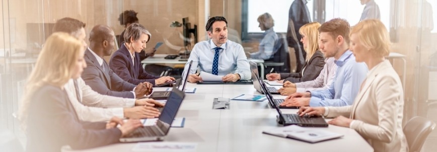 A group of business professionals sit around a conference table in a meeting room, using laptops and taking notes, while a man at the head of the table discusses the company’s risk management plan with the group.