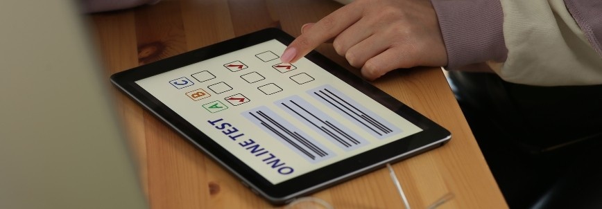 A person is using a tablet on a wooden desk to take an online test, selecting answers from multiple-choice options displayed on the screen, similar to the CAPM exam format.