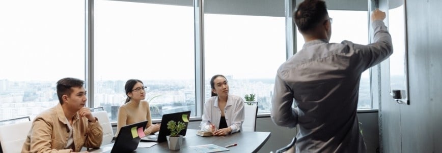 A man presents at a whiteboard to three colleagues in a modern office with large windows. They listen attentively with laptops and papers, discussing CAPM job opportunities in the current business landscape.