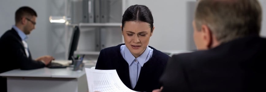 A woman sitting at a desk looks upset while holding documents, possibly due to PMP exam mistakes, as she faces a man who is talking to her. Another man works at a computer in the tense office environment.