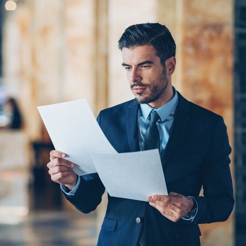 A man in a dark suit and tie stands indoors, holding and reading two sheets of paper with expert judgment instructions, his expression focused. The background is blurred with warm tones.
