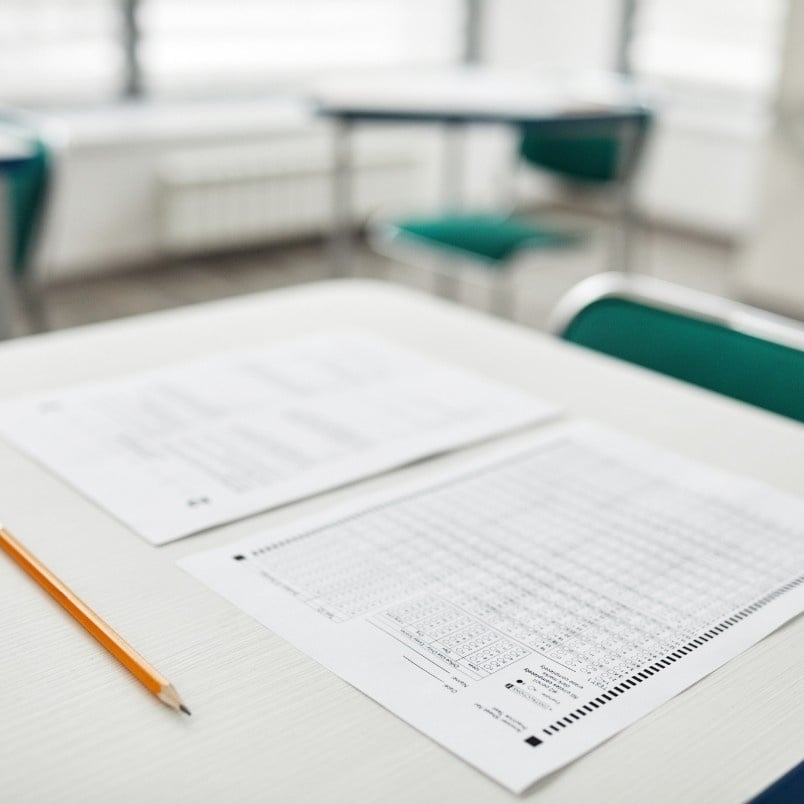 A close-up of a standardized test answer sheet and a pencil on a white desk in a classroom, suggesting students preparing with CAPM exam practice questions; empty green chairs and more desks are blurred in the background.
