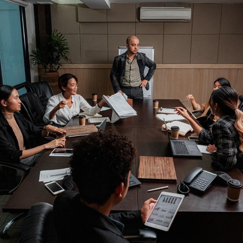 Six people sit around a conference table with laptops and documents, listening as one person stands at the head of the table, providing retrospective instructions during an engaging business meeting.