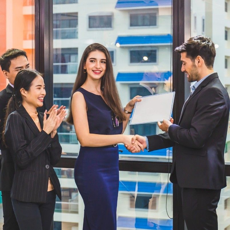 A man in a suit hands a CAPM certification to a smiling woman in a blue dress as they shake hands. Two colleagues beside them, a man and a woman, clap and smile. They are indoors with large windows showing buildings outside.