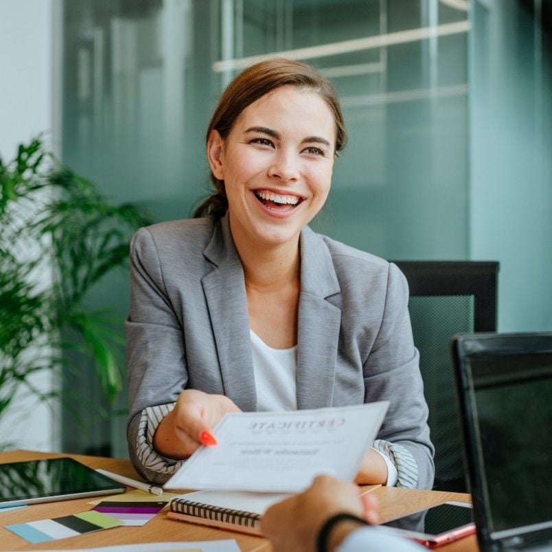 A woman in a gray blazer smiles brightly while handing a certificate across a desk in an office setting—perhaps celebrating success after overcoming common PMP exam mistakes. Papers, a laptop, and green plants are visible in the background.