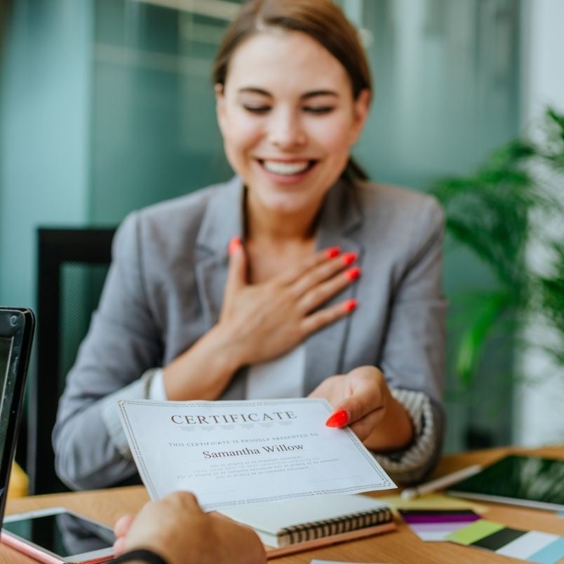 A smiling woman in a blazer receives a certificate while placing her hand on her chest, showing appreciation—perhaps for meeting PMP certification requirements. She sits at a desk with a laptop, tablet, and plant in the background.