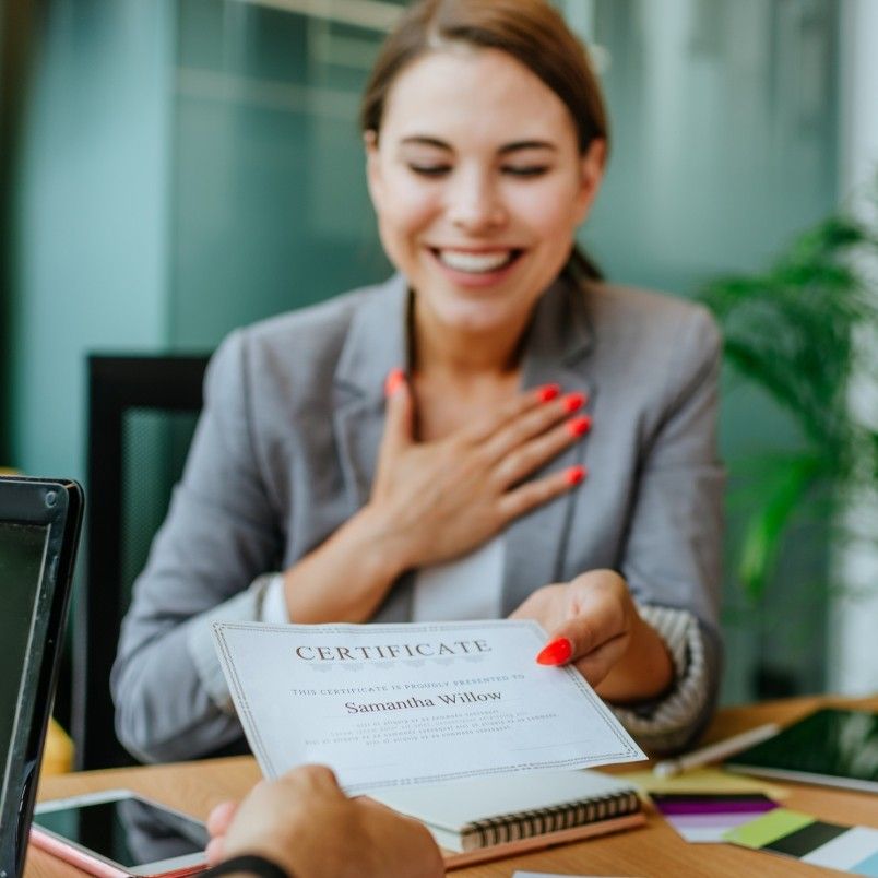 A smiling woman in a blazer sits at a desk, proudly holding out her CAPM certification with one hand while her other hand rests on her chest, looking pleased and grateful.