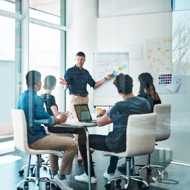 A man stands and gestures while presenting a scope statement to four seated colleagues in a modern glass-walled office, with charts and sticky notes visible on the whiteboard and walls.