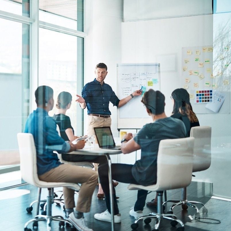 A man stands by a whiteboard, using an epic template to give a presentation to four colleagues seated around a conference table in a modern office with large windows. Notebooks and laptops are on the table.