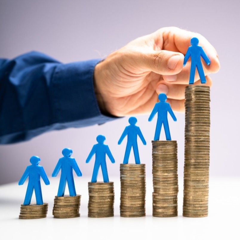 A hand places a blue paper figure on the tallest stack of coins, with others on shorter stacks, symbolizing financial growth or career progression—much like how earning a CAPM certification can boost your salary prospects.