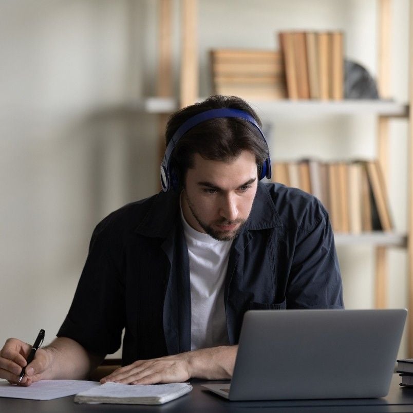 A man wearing headphones sits at a desk, focused on his laptop while researching if CAPM Certification is worth it, taking notes on paper, with bookshelves in the background.