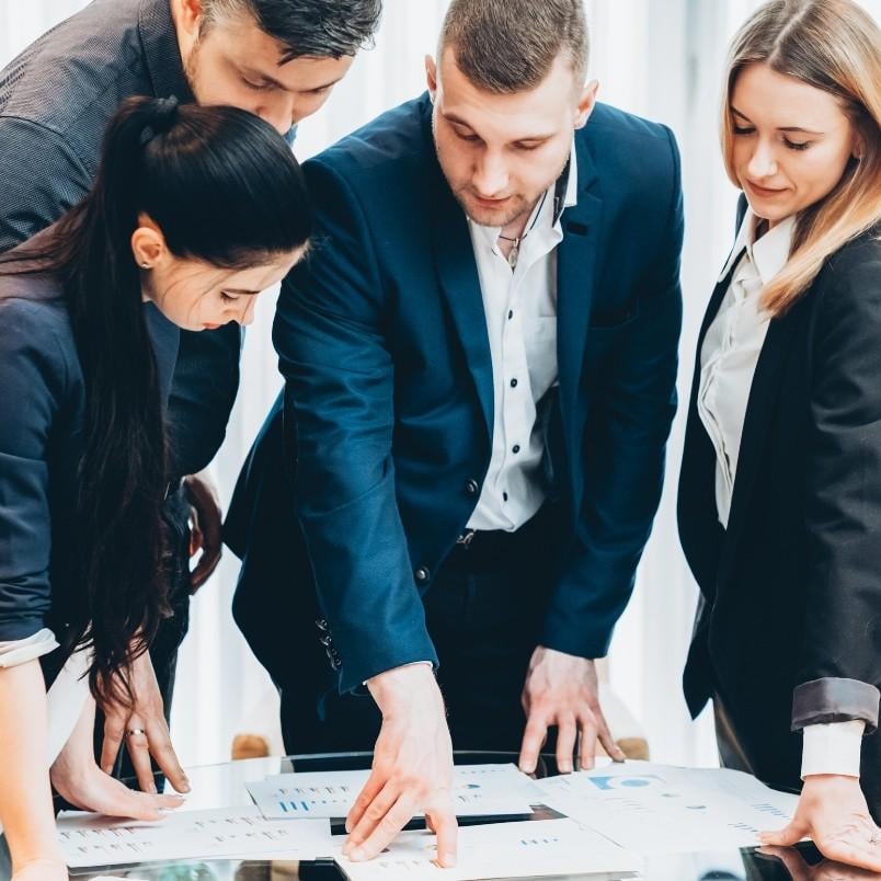 Four business professionals, dressed in formal attire, are gathered around a table reviewing charts and documents, appearing focused and engaged in discussion about their procurement management plan.