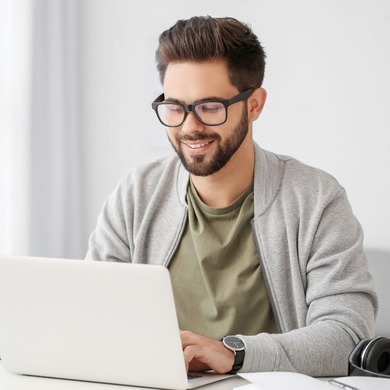 A smiling man with dark hair, beard, and glasses sits at a desk using his laptop for a CAPM Exam Simulation. He wears a gray jacket over an olive green shirt. Headphones and a pen rest on the desk in the bright, minimalistic background.