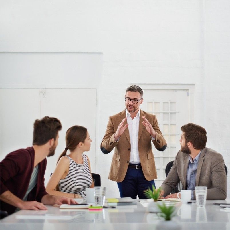 A man in a brown blazer stands and speaks to three seated colleagues at a table in a modern office, gesturing with his hands as they use the nominal group technique to encourage attentive listening and idea sharing.