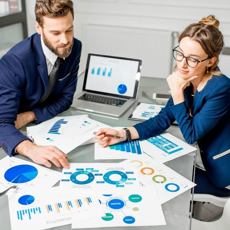 Two business professionals sit at a glass desk reviewing printed charts and graphs, with a laptop displaying data visualizations and product roadmap instructions open in the background. Both appear focused and are dressed in business attire.