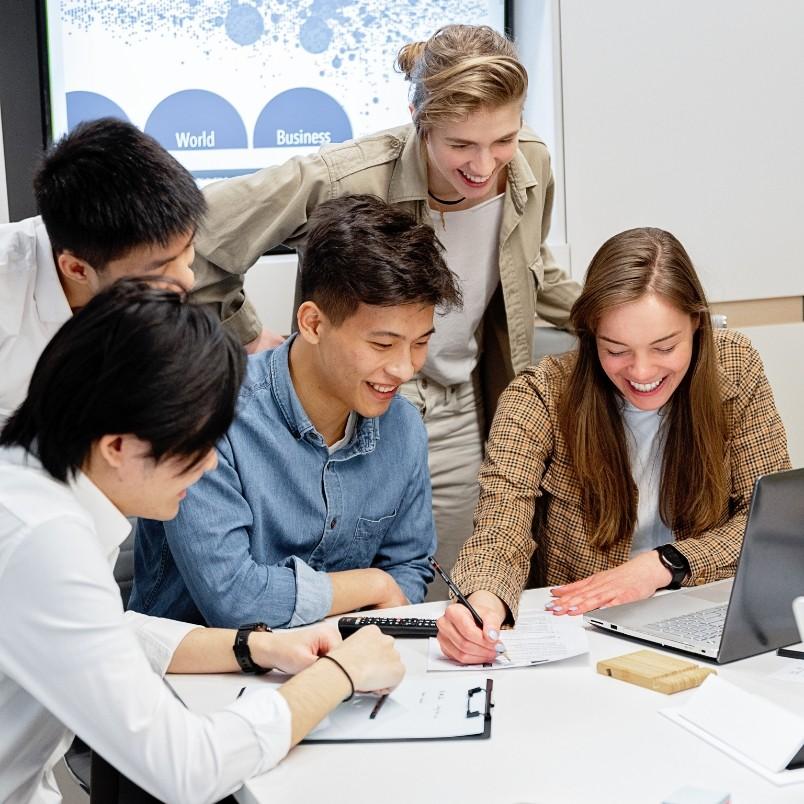 Five young adults gather around a table, smiling and collaborating on a procurement management plan with a laptop, clipboard, and papers, creating a positive and engaged group atmosphere in a modern office setting.