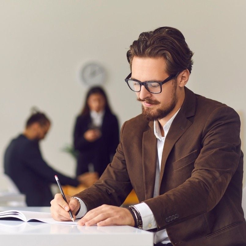 A man with glasses and a brown blazer sits at a desk, conducting Checklist Analysis while writing with a pencil. In the blurred background, two people are talking and another is sitting, working on a laptop.