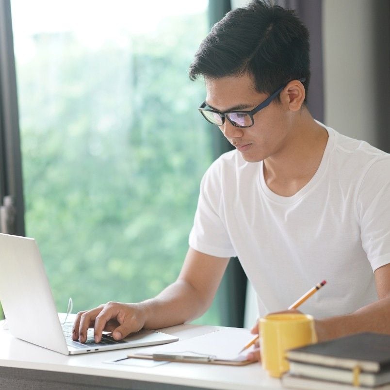 A young man wearing glasses and a white t-shirt works on a laptop at a desk by a window, jotting down notes about PMP exam mistakes in his notebook. Books and a cup sit nearby, while greenery is visible outside.