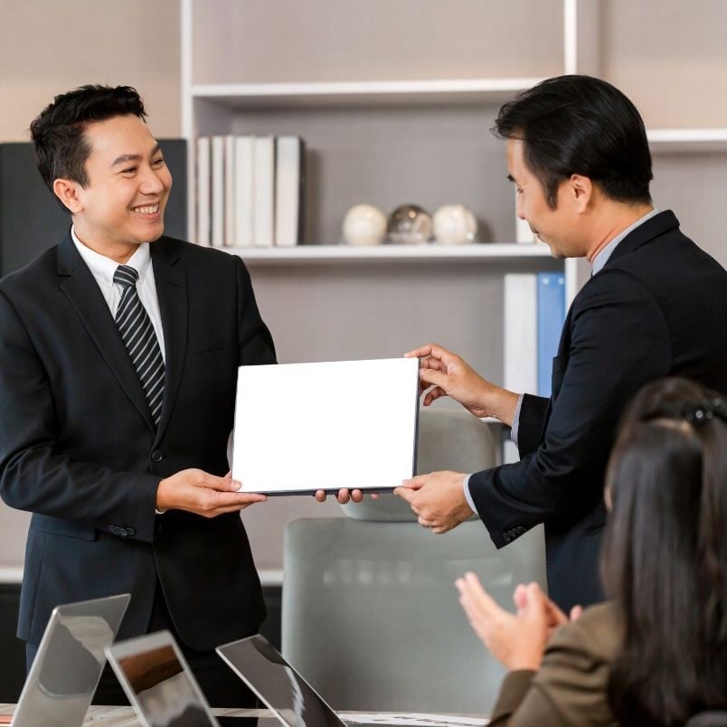 Two men in business suits stand in an office, smiling as one hands a blank certificate to the other—perhaps celebrating meeting PMP certification requirements. Colleagues seated at a table with laptops applaud in the foreground.