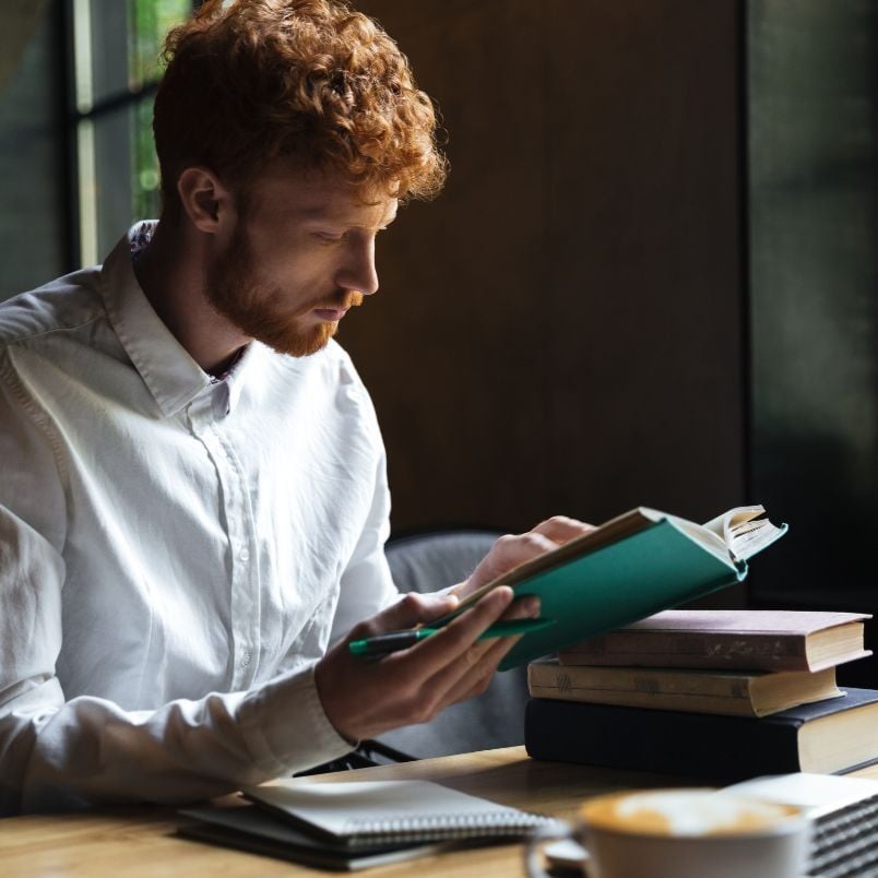 A young man with curly red hair and a beard reads about PMP certification requirements at a wooden table, with notebooks and stacked books nearby. He is wearing a white shirt and is focused on his reading in a softly lit room.