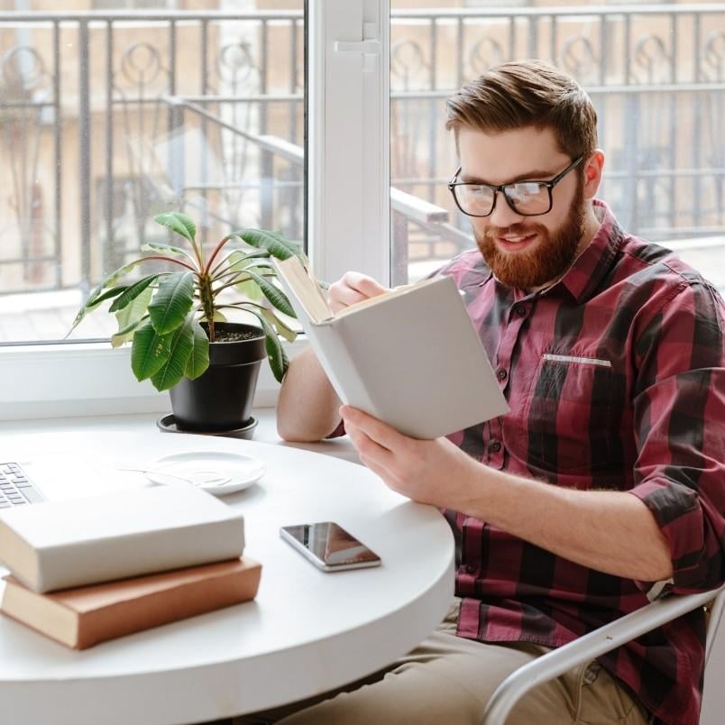 A man with a beard and glasses, wearing a red and black plaid shirt, sits at a round table by a window reading a book, possibly preparing for the CAPM Exam Simulation. Other books, a smartphone, laptop, and potted plant are on the table.