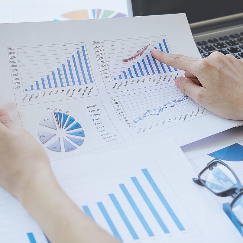 Close-up of hands holding and pointing at printed business charts and graphs, including bar graphs, a pie chart, and a line graph, performing Root Cause Analysis, with a laptop and eyeglasses in the background.