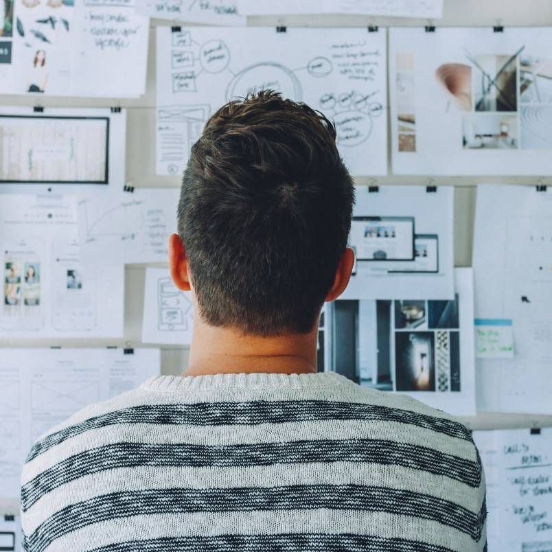 A person with short brown hair and a striped sweater is seen from behind, looking at a wall covered with papers, diagrams, and photographs—perhaps reviewing a scope statement or planning their next move.