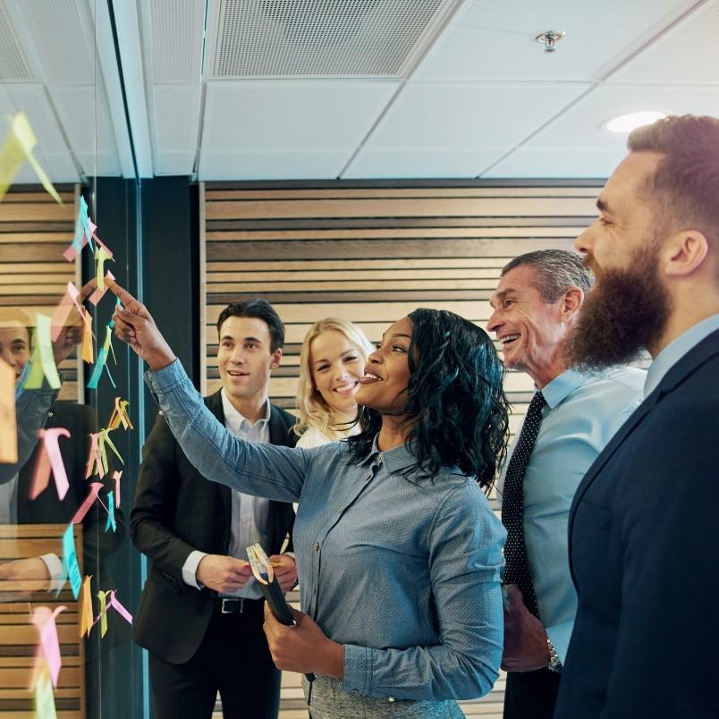 A group of four professionally dressed people smile and collaborate, enhancing PROJECT COMMUNICATION as they place colorful sticky notes on a glass wall in a modern office setting.