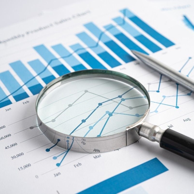 A magnifying glass and pen rest on printed financial charts, including blue bar graphs, line graphs, and risk burndown chart instructions highlighting monthly data trends and key figures.