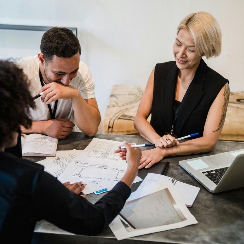 Three people sit around a table with papers, notebooks, and a laptop, smiling as they review product roadmap instructions while collaborating in a casual office setting.