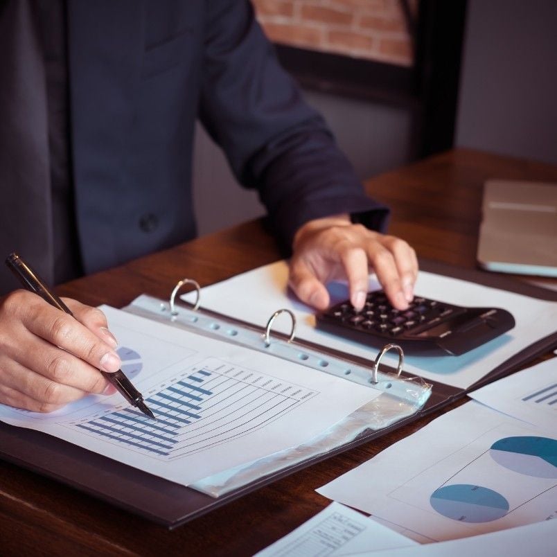 A person in business attire conducts a checklist analysis, reviewing a financial chart in a binder, holding a pen in one hand and using a calculator with the other on a wooden desk with documents and a laptop.