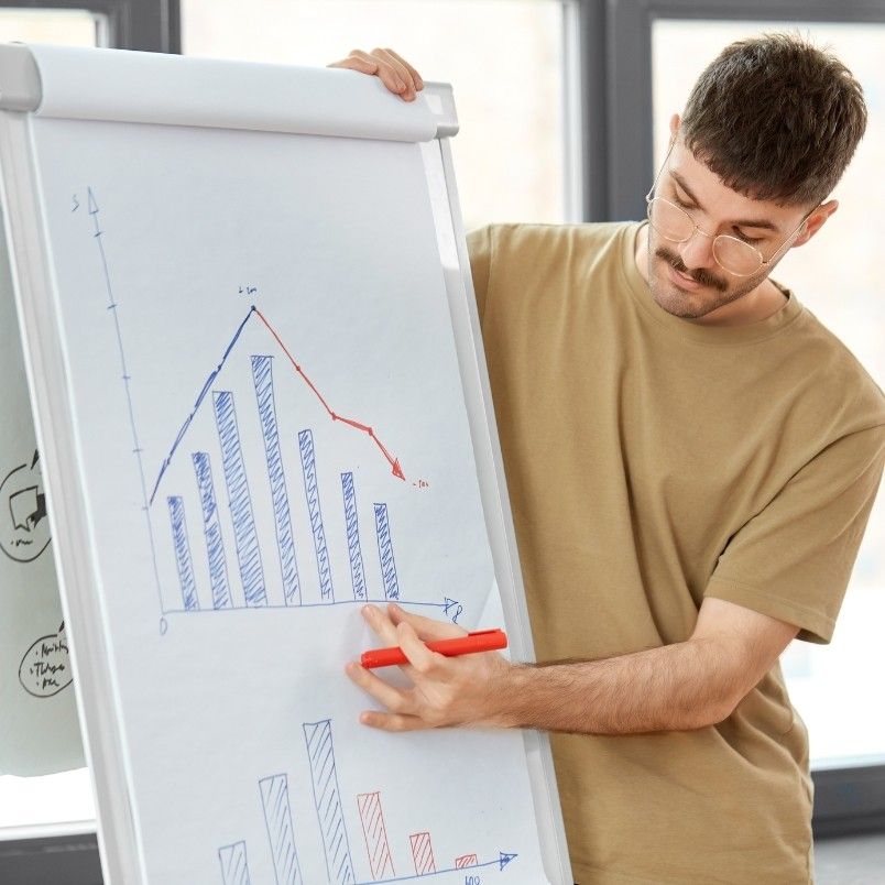 A person wearing glasses and a beige t-shirt points to a bar and line graph on a flip chart, providing risk burndown chart instructions while explaining data trends in an office setting.
