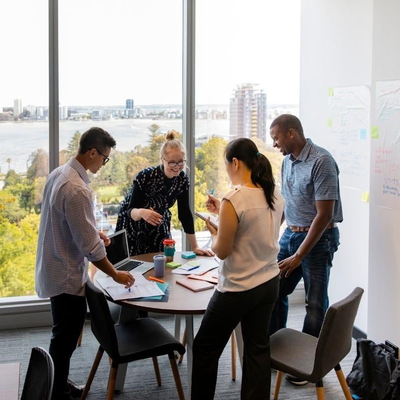 Four people stand around a round table in a modern office with large windows overlooking a city and river, engaging in project communication as they collaborate on documents and laptops, with whiteboards and notes visible.