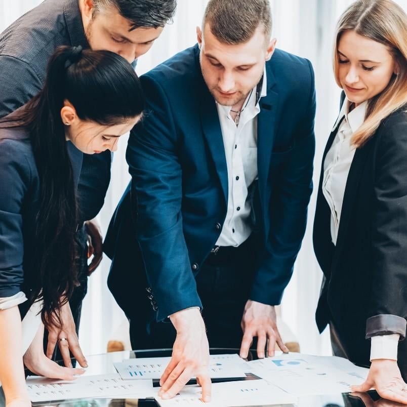 Four business professionals, two men and two women, stand around a table in a bright office reviewing charts, graphs, and product roadmap instructions as they collaborate and discuss key data.
