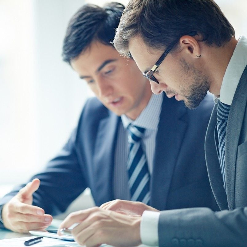 Two men in business suits sit closely together, focused on documents and a tablet in front of them. One points at the tablet while the other observes, both appearing engaged in a discussion or review of retrospective instructions.