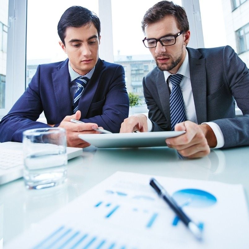 Two businessmen in suits sit at a desk reviewing a scope statement on a tablet. A glass of water, pen, and printed charts with graphs are on the table. Large windows provide a bright background.