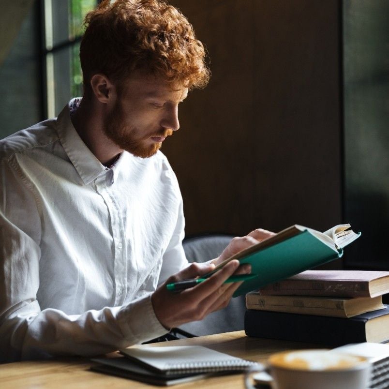 A young man with curly red hair and a white shirt reads a book on how to pass the CAPM exam at a wooden table with a notebook, pen, and stacked books. Soft natural light comes through a window in the background.