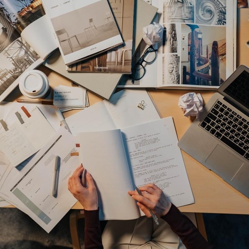 A person sits at a cluttered desk with open notebooks, magazines, a laptop, pens, coffee, and crumpled paper, appearing to study hard on how to pass the CAPM exam or work on architectural or design projects.