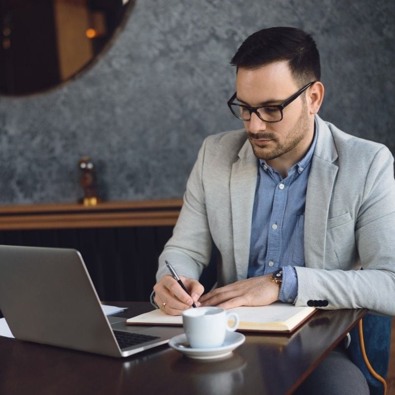 A man wearing glasses and a light gray blazer sits at a table with a laptop, notebook, and cup of coffee, writing study notes on how to pass CAPM exam while working in a modern, stylish café.