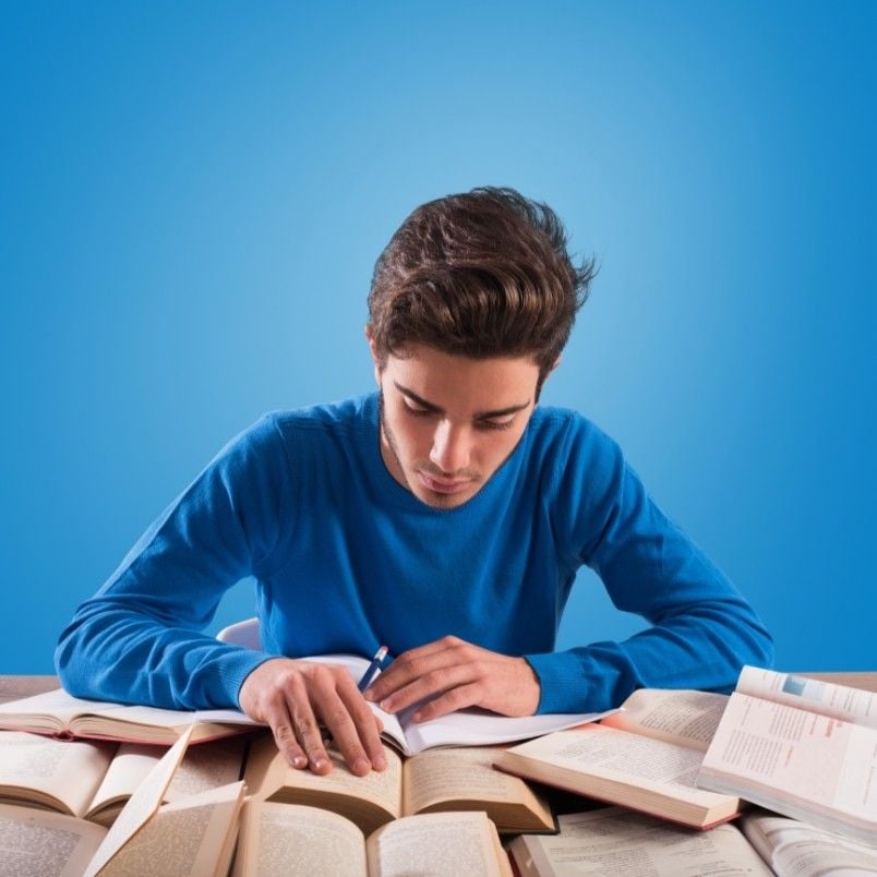 A young person in a blue shirt is sitting at a table, surrounded by open books, focused on studying and taking notes on how to pass the CAPM exam, with a blue background behind them.