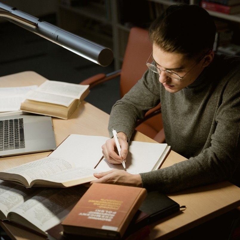 A person with glasses sits at a desk in a library, writing in a notebook on how to pass the CAPM exam. Open books and a laptop are on the table, and a lamp illuminates the workspace.