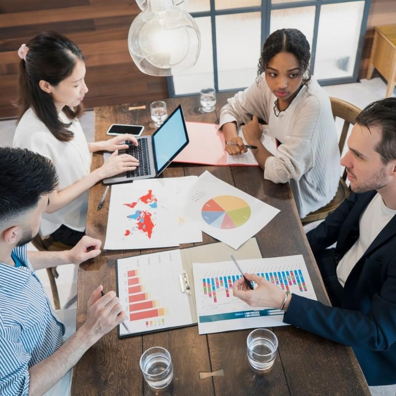 Four people sit around a table with laptops, charts, and documents, following expert judgment instructions during a business meeting. Colorful graphs and a world map are visible on the table, along with glasses of water.
