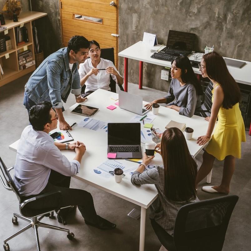 Six people in an office are gathered around a table with laptops, documents, and coffee cups, actively discussing expert judgment and reviewing instructions during a collaborative meeting.