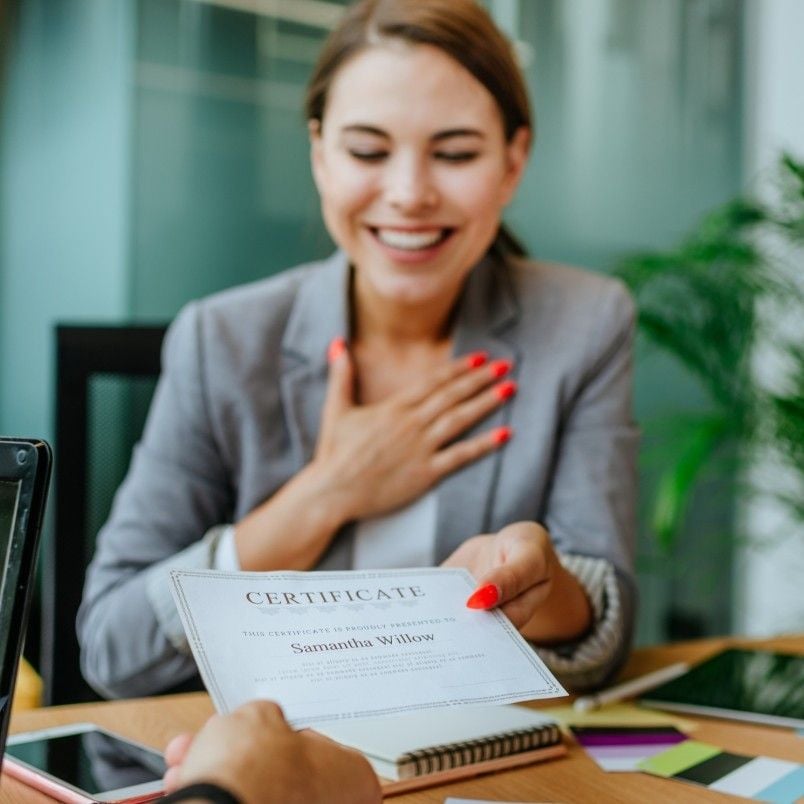 A smiling woman in a gray blazer sits at a desk, placing one hand on her chest and handing a certificate to another person. She appears happy and appreciative, perhaps celebrating after overcoming the setback of failing the CAPM exam.