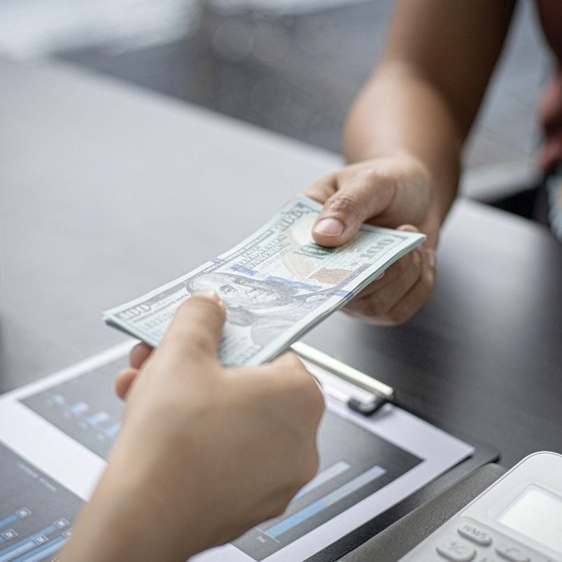 A person hands a stack of U.S. hundred-dollar bills to another across a desk with financial documents and a calculator, highlighting the question: Is CAPM Certification worth it for your financial future?.