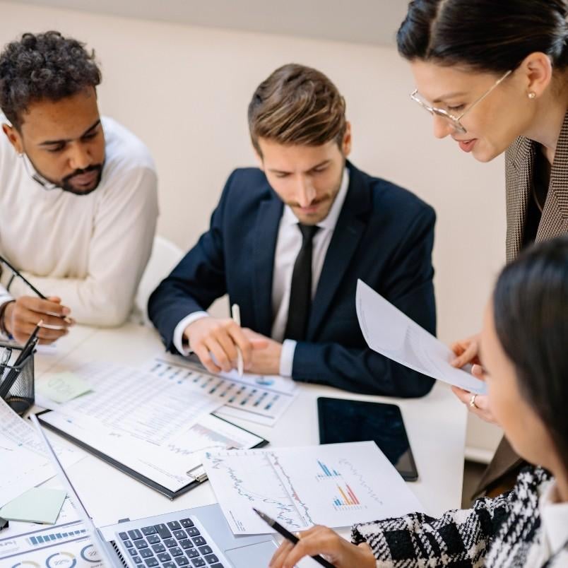 Four professionals collaborate at a desk covered with charts, graphs, and a laptop, using expert judgment to interpret instructions and discuss documents and financial data in a modern office setting.