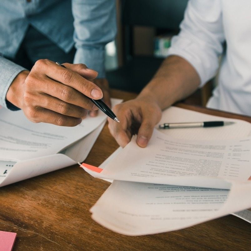 Two people are reviewing and discussing documents at a desk, possibly working through CAPM exam practice questions. One person points at a paper with a pen, while the other's hand rests nearby amid papers, folders, and a notebook spread across the table.